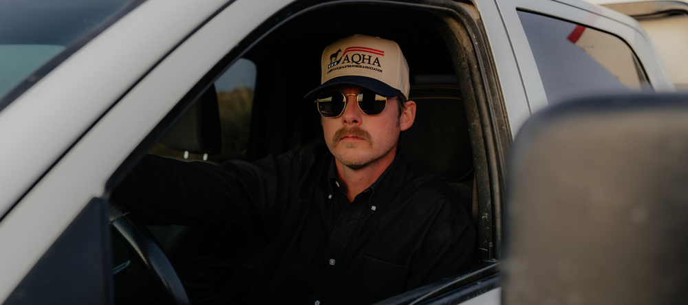 Man sitting in a vehicle wearing a cap and sunglasses, with a blurred background